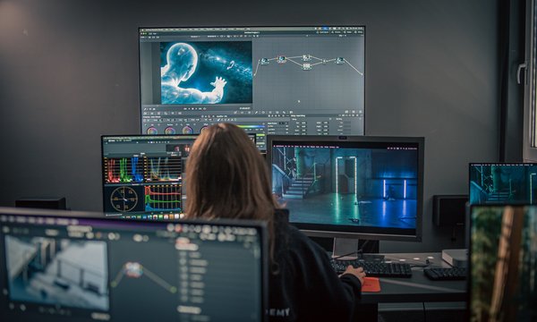 W słabo oświetlonym pomieszczeniu osoba siedzi przy biurku, na którym stoi kilka monitorów wyświetlających oprogramowanie do montażu wideo, narzędzia do korekcji kolorów oraz futurystyczną grafikę cyfrową.